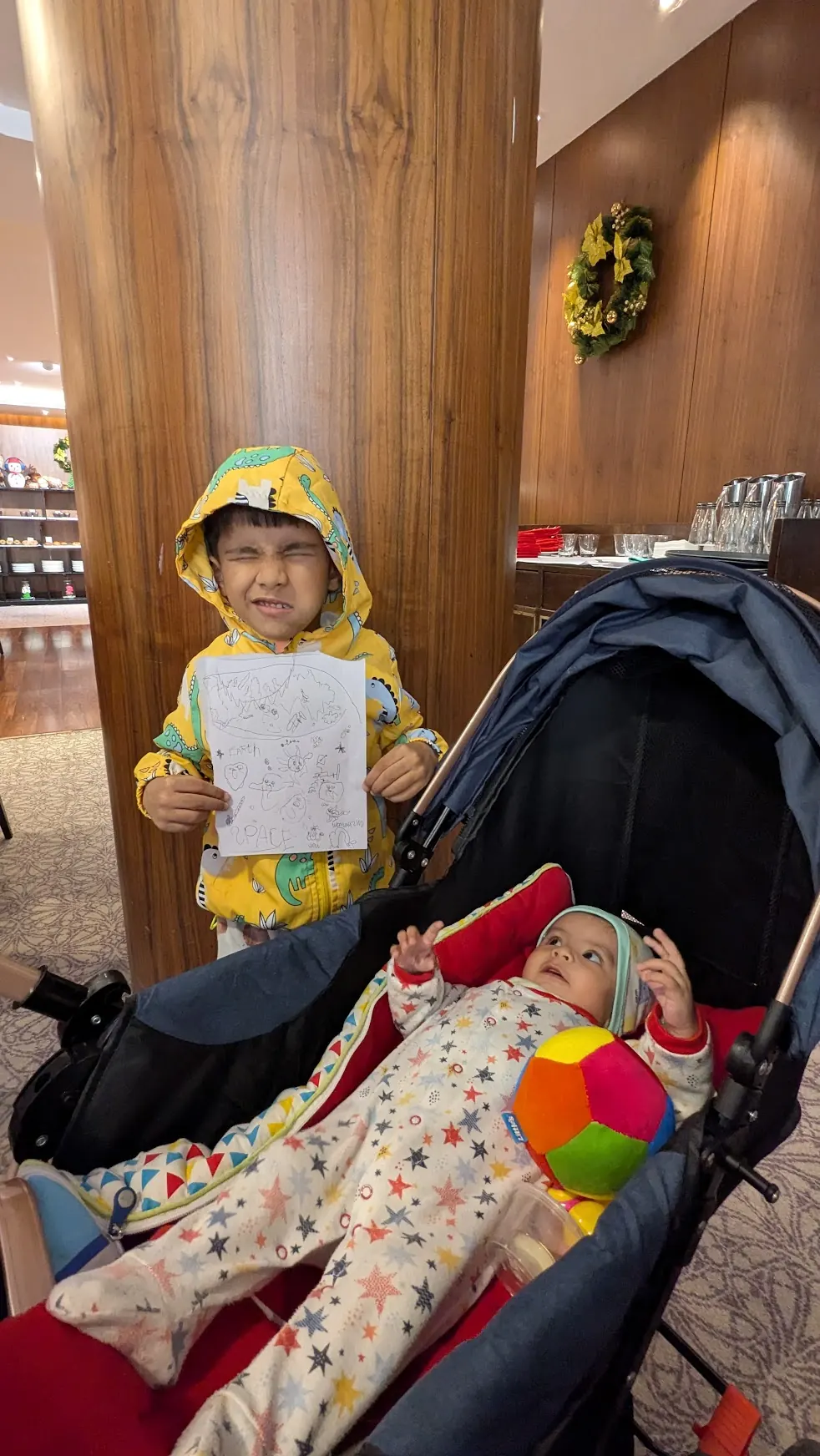 A boy holding up a drawing beside a baby in a stroller